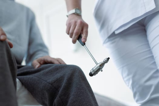 Doctor testing patient knee reflex using neurological hammer during physical examination in clinic.