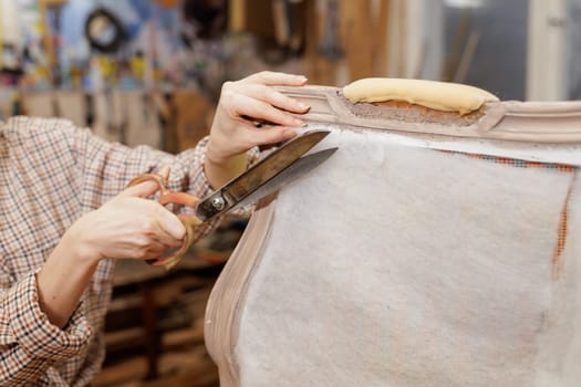 Close-up of hands cutting upholstery fabric with large scissors while restoring vintage furniture in a workshop.