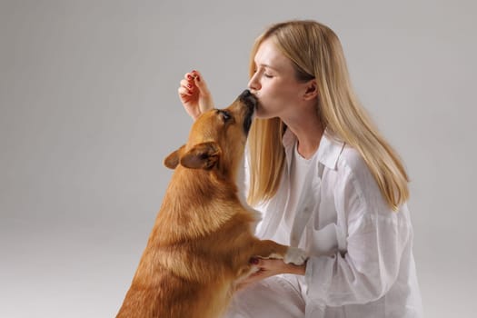 Happy woman kisses her adorable Welsh Corgi dog in studio, showing affection and strong human-animal bond.