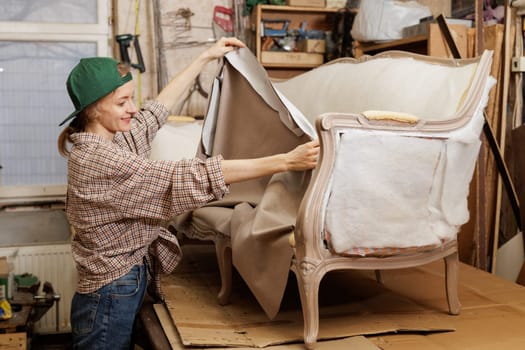 Smiling female upholsterer stretching fabric over vintage sofa frame while restoring furniture in workshop.