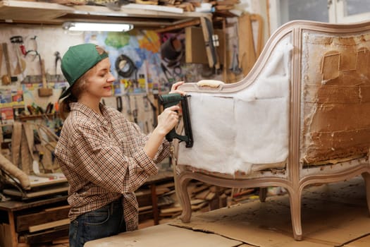 Smiling woman upholstering antique armchair with pneumatic stapler in workshop, engaged in restoration work.