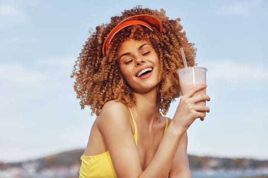 A cheerful woman with curly hair holds a refreshing beverage, wearing a visor, set against a sunny backdrop