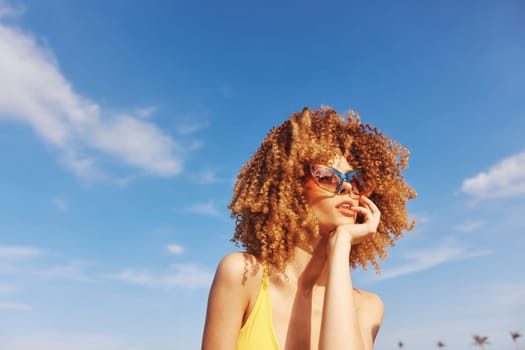 A fashionable young woman with curly hair and colorful sunglasses, relaxing outdoors under a bright blue sky with fluffy clouds