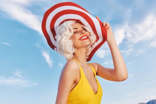 A cheerful woman poses playfully in a vibrant yellow swimsuit with a stylish red and white hat under a sunny sky
