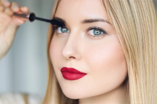 Woman applying mascara with confidence, defined brows and red lipstick. Blonde beauty focused on her eye makeup, cosmetics look