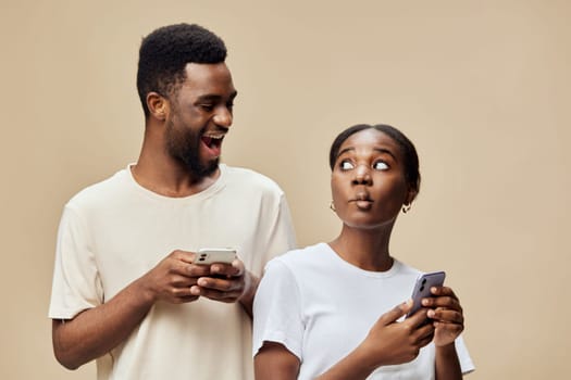 A joyful Black couple interacts with their smartphones, expressing surprise and curiosity, set against a soft beige background, ideal for showcasing modern relationships