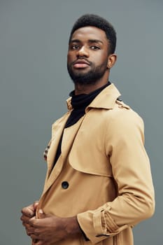 An elegant African man dressed in a beige trench coat stands against a neutral gray backdrop, exuding confidence and modern style The focus is on contemporary fashion, emotions, and self expression