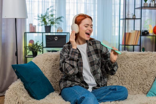 Young woman at home wearing headphones singing joyfully using comb as microphone and enjoying music fun. Caucasian redhead girl on sofa smiling having playful emotional moment with creative expression