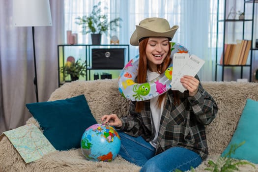 Young woman at home with inflatable swim ring looking at globe and holding plane tickets planning vacation. Caucasian redhead girl on sofa dreaming about travel, smiling with excitement and wanderlust