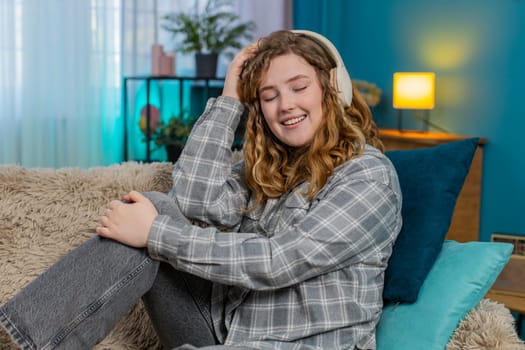 Young woman at home wearing headphones with closed eyes gently dancing and enjoying favorite music. Caucasian redhead girl on sofa moving to rhythm, feeling joy, emotional peace and relaxation