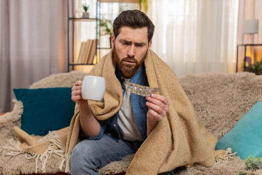 Young man at home sits on the sofa, wrapped in a warm blanket, holding a cup of tea to feel better. Caucasian guy looks unwell, with medicine on the table, possibly recovering from illness or cold