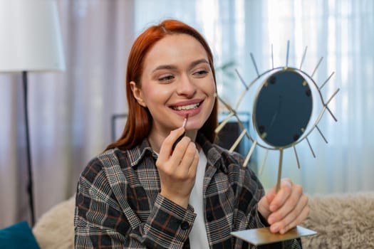 Young woman at home applying makeup with focus, drawing lips using lipstick and smiling into mirror. Caucasian redhead girl on sofa enjoying beauty routine with care, confidence and joyful emotion