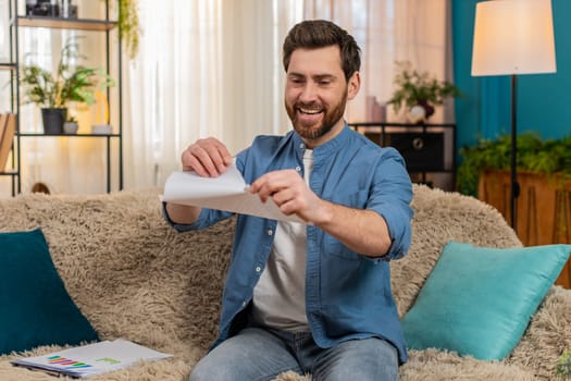 Young man at home sits on the sofa, smiling and tearing an unnecessary document into pieces with relief. Caucasian guy feels happy and free, no longer worried about obligations, debts, or past issues