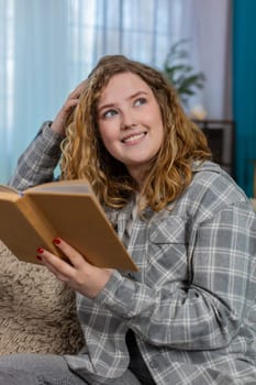 Young woman at home reading book with focus and flipping pages slowly while reflecting on deep content. Caucasian redhead girl on sofa engaged in thoughtful reading, enjoying quiet intellectual moment