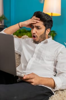 Indian man feeling frustrated at home, reacting to online gaming loss, showing disappointment and tension. Arabian guy sitting on sofa, stressed after failed gaming attempt, focused on laptop screen