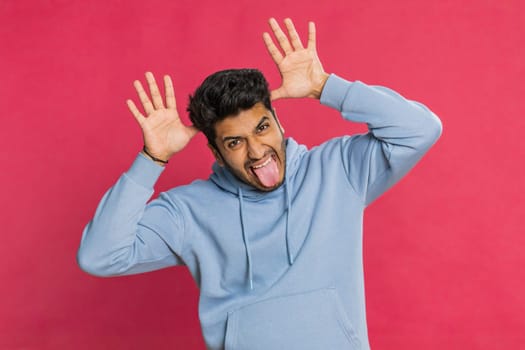Funny comical playful Indian man making silly facial expressions, grimacing, fooling around, showing tongue, idiotic expression. Comedian Arabian guy isolated on red background. People lifestyles