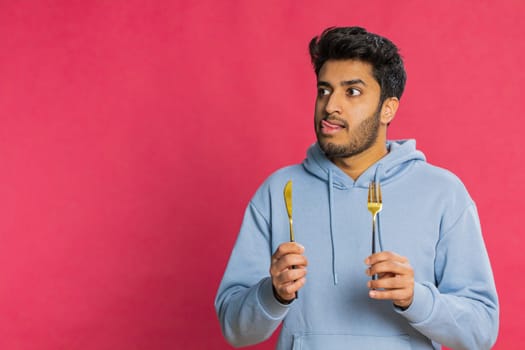 Hungry Indian man holding fork and knife, eagerly anticipating a delicious meal, licking lips with excitement. Starving Arabian guy craving food with impatient expression isolated on red background