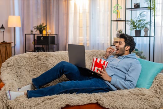 Indian young man eating popcorn and watching movie on laptop, enjoying relaxed time. Hispanic guy sits on sofa in living room smiling while having snacks and staying entertained with favorite film.