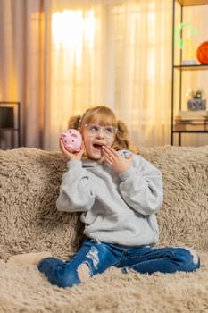 Young teenager girl holds a toy piggy bank at home, rattling it gently to hear the metallic coins clink. Child kid on sofa smiles broadly, thrilled excited about setting save money for a dream item.