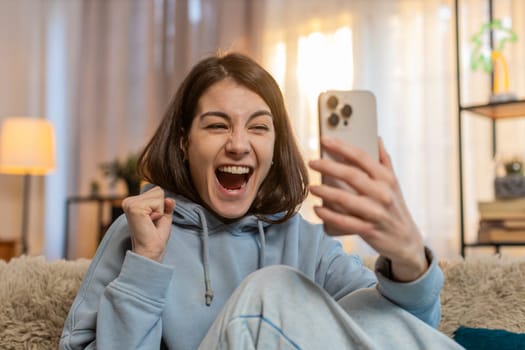 Excited Caucasian young woman using smartphone typing browsing found out great win in game or good news celebrate victory. Girl with mobile phone happily jumping and shouting with joy sitting on sofa.