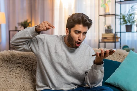 Angry mad stressed Indian young man arguing while talking on smartphone via speaker mode irritated shout quarrel. Hispanic offended dissatisfied guy in casuals sitting on sofa in living room at home.