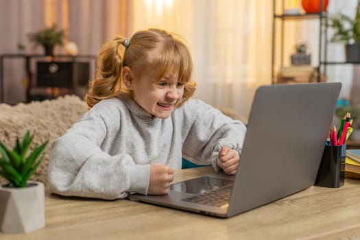 Young girl sitting at a table with a laptop, feeling frustrated after losing a game at home. Caucasian child showing disappointment with an upset expression, reflecting emotions of failure and sadness