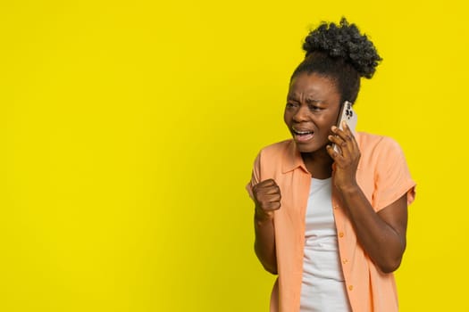 African American woman talking on phone, gesturing angrily expressing frustration displeasure quarrel. Black girl isolated on yellow background negative emotions and tension during heated conversation