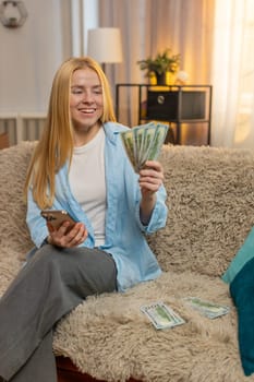 Young woman relaxes at home, counting banknotes while entering sums on smartphone to find extra savings. Caucasian girl on sofa celebrates with triumph pose, feeling proud of unexpected financial gain