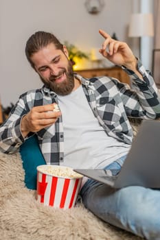 Young man eating popcorn at home, occasionally tossing kernels playfully while watching funny clips on laptop. Caucasian guy relaxes comfortably on sofa, laughing out loud at silly and amusing videos