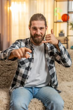 Young man sitting at home sofa with hand shaped like a phone, smiling in open anticipation for future call gesture asking for conversation. Caucasian guy waiting for contact from someone important.