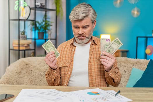 Mature man sitting at home table, surrounded by documents bills, holding a small amount of money dollars cash. Guy exhausted disappointed due to financial difficulties bankruptcy, stress frustration