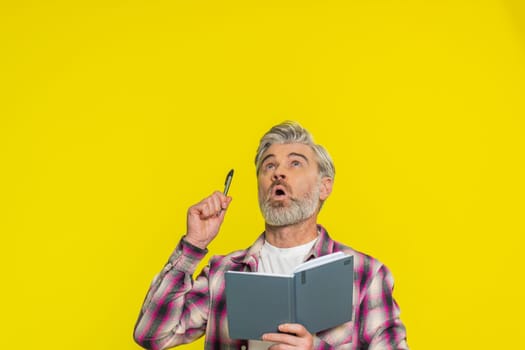 Thoughtful journalist Caucasian middle-aged man making notes, writing down thoughts with pen into notepad notebook diary, to do list, good idea. Mature old guy isolated on yellow background indoors