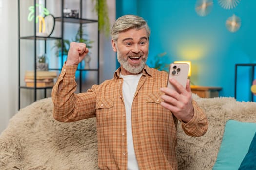 Caucasian mature man sitting on sofa at home, smiling joyfully while holding smartphone. Middle-aged guy celebrates successful project completion online game victory, expressing satisfaction happiness