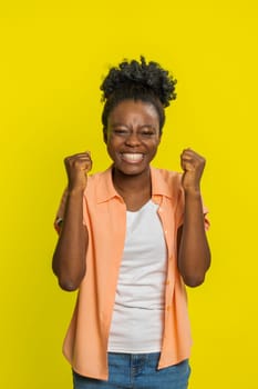 Happy joyful African American woman celebrating success victory, winning birthday lottery jackpot goal achievement play game good positive news triumph. Young Black girl isolated on yellow background