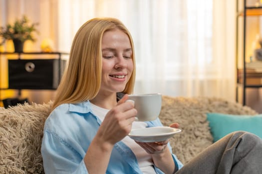 Woman tasting drinking warm coffee at home, closing eyes in mild bliss while taking a slow sip. Girl sitting at table on sofa regains vitality blending quiet enjoyment with positive early morning mood