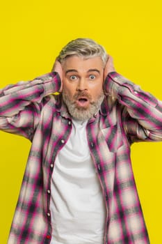 Mature man covers ears in strong panic, showing massive fear, hesitation and alarmed reaction to huge chaos. Middle-aged guy on yellow background indicates intense shock from unexpected loud noise.