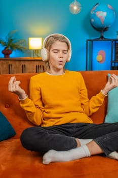 Caucasian teenager boy meditates in lotus pose on comfortable sofa at home. Wearing headphones listening music, child relaxes unwinds peacefully after long school day, finding inner peace calmness
