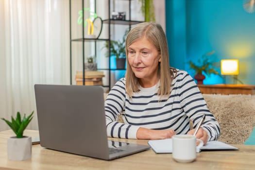 Mature woman participating in online course on laptop. Caucasian grandmother taking notes while sitting on sofa at home table, engaging in learning activities enhancing knowledge, education lessons