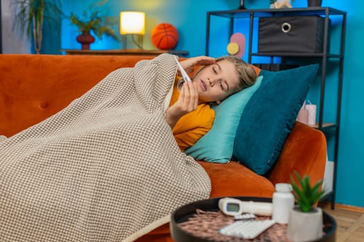 Caucasian teenager boy is sick at home. Lying on sofa with thermometer, checks high temperature, sad and tired, medicine packages nearby, displaying emotions of illness and fatigue in a living room