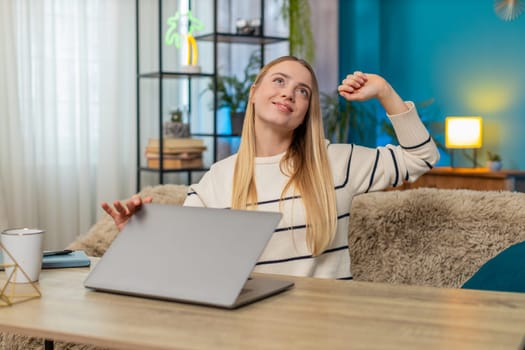 Caucasian woman sits at home on table finishing a remote workday on laptop, stretching, smiling, feeling calm and fully satisfied. Girl appears happy, relieved, enjoying calm work, continuing effort