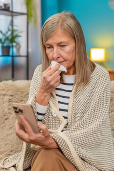 Caucasian ill mature woman sits on home sofa covered with blanket, looking tired upset holding smartphone. Sick grandmother tries to order medicine online, expressing frustration exhaustion, cold flu