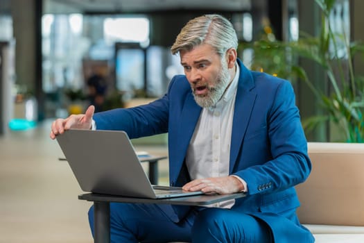 Sad middle-aged Caucasian businessman using laptop typing browsing, loses becoming surprised sudden lottery results, bad news, fortune loss fails office lobby. Upset freelance worker in hotel hall.