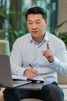 Young Chinese businessman writing down or taking down notes while attending online video class or office meeting on laptop in hotel lobby. Confident manager in shirt support services looking at webcam