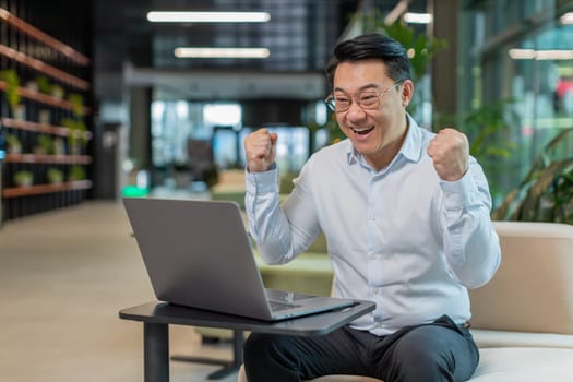 Chinese young businessman working on laptop shocked by sudden victory, game winning lottery goal achievement, good career news. Excited guy freelancer clenching fists sitting in modern office lobby.