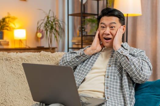 Surprised Chinese middle-aged man using laptop, receive good news shocked by sudden victory celebrate lottery jackpot win at home apartment. Cheerful guy with netbook sitting on sofa in living room.