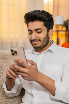 Indian young man at home sitting on sofa focused on smartphone typing text, browsing news, reacting to notifications. Arabian guy using mobile phone for social media, work, texting, digital lifestyle
