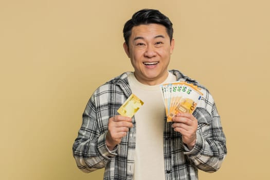 Happy Asian middle-aged man showing plastic credit bank card and money euro cash advertising taking loan financial help for purchases online. Mature Chinese guy isolated on beige background indoors