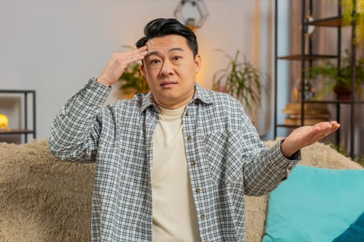 I dont know what to say. Confused Asian handsome man feeling embarrassed about ambiguous question, having doubts, no idea, being clueless and uncertain at home. Chinese guy sits on sofa in living room