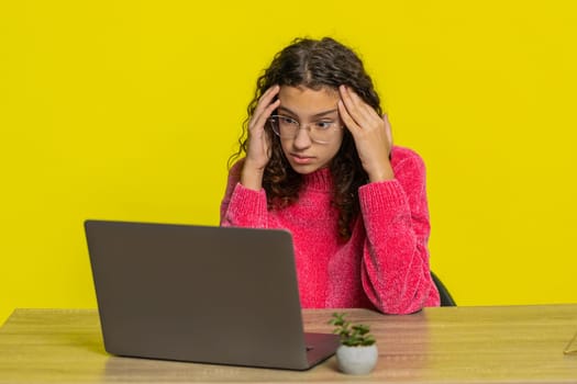 Displeased young adult girl using laptop notebook typing working, loses becoming surprised sudden lottery results bad news fortune loss game fail computer virus sitting at table on yellow background.