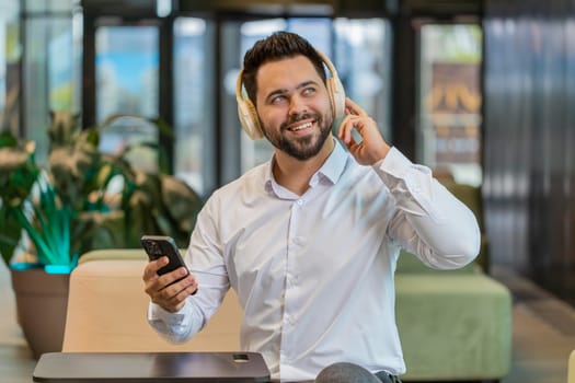 Happy relaxed Caucasian young businessman wearing headphones listening favorite energetic disco music on smartphone. Male entrepreneur in formal relaxing, taking a break sitting in modern office lobby